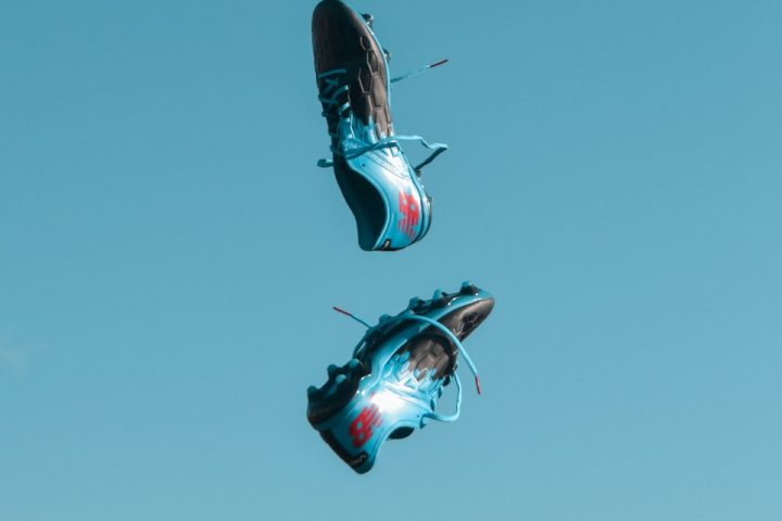 pair of blue-and-black New Balance cleats in the air under teal sky with white clouds